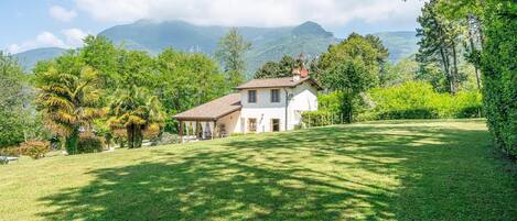 A house with a tiled roof, surrounded by a well-maintained lawn and trees, with mountains in the background.