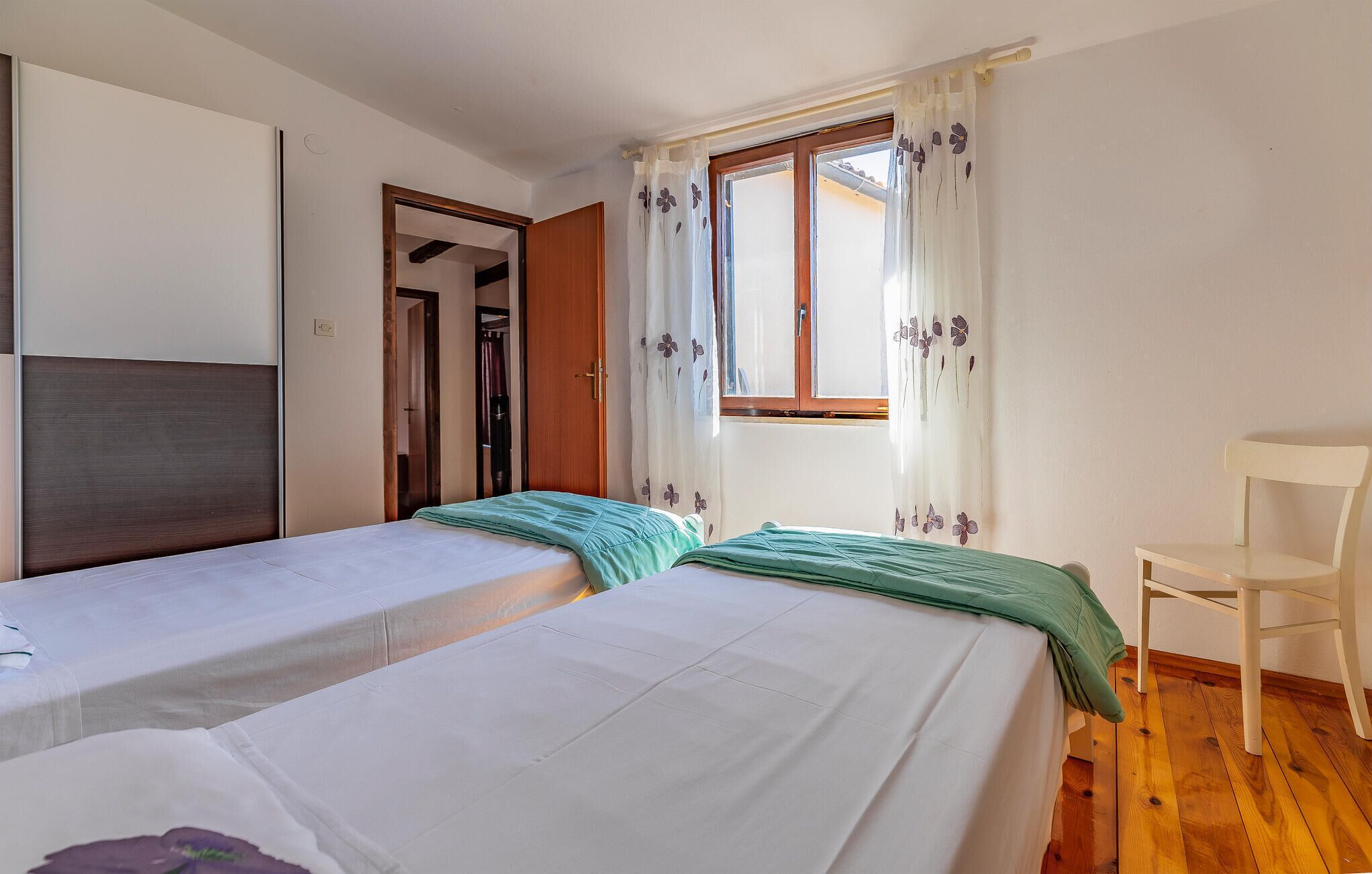A bedroom with two single beds, a wooden floor, a white chair, and a window with floral curtains.