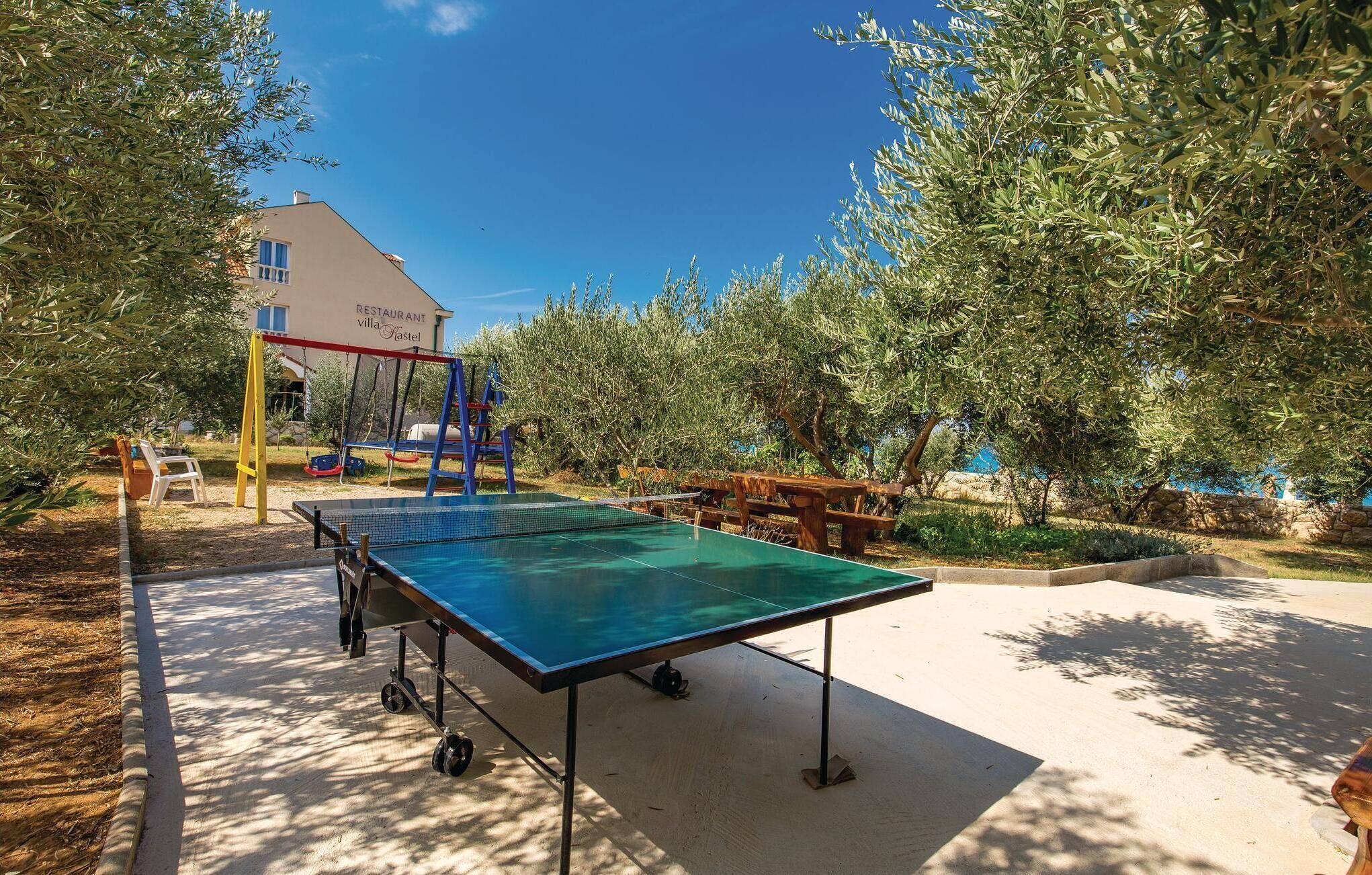 A ping pong table in an outdoor area with a swing set and a building labeled "RESTAURANT villa Fastal" in the background.