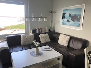 Uma sala de estar com um sofá de couro marrom, almofadas brancas e fofas, uma mesa de centro branca com um vaso de flores, um quadro na parede e uma estante de livros com livros e um rádio.
