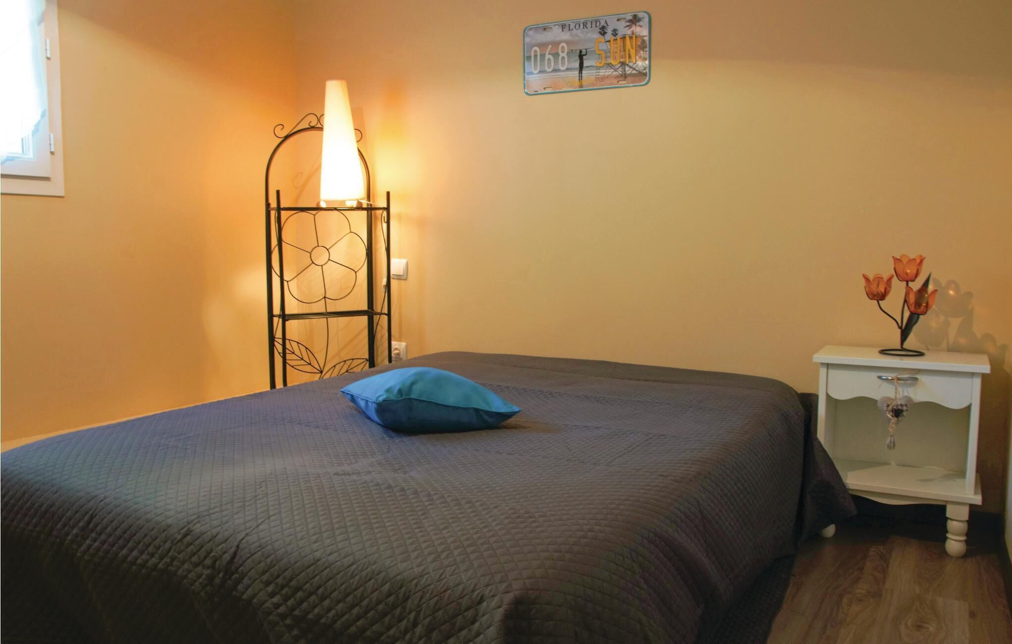 Une chambre à coucher avec un lit, une table de chevet ornée d’un vase de fleurs, une horloge murale et un tableau représentant une scène de plage.