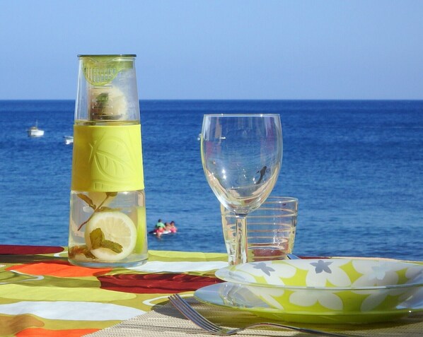 A table set with a glass bottle, a wine glass, and a bowl, with a view of the sea and boats in the background.
