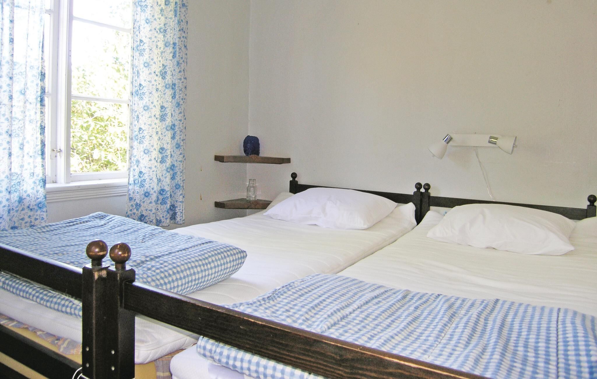 A room with two single beds, a window with blue floral curtains, a bedside table with a lamp, and a small shelf with a blue object.