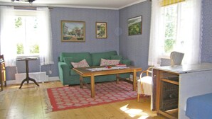 Ett vardagsrum med en grön soffa, ett soffbord i trä och en röd, mönstrad matta. På väggen hänger två inramade tavlor och det finns ett fönster med gardiner.