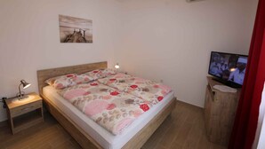 A bedroom with a wooden bed, a nightstand with a lamp, a television on a wooden stand, and a painting on the wall. - Holiday apartment with air conditioning (Šilo)