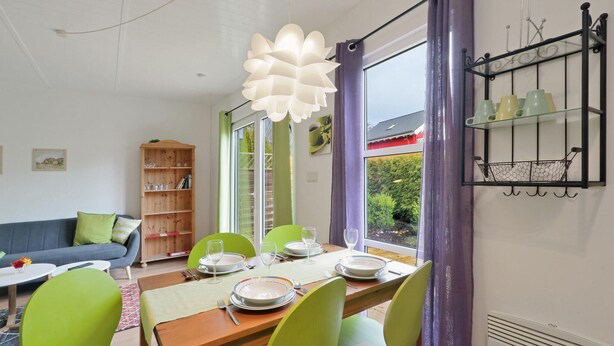 Un comedor con una mesa de madera dispuesta para cuatro personas, rodeado de sillas verdes, y un ventanal con cortinas moradas.