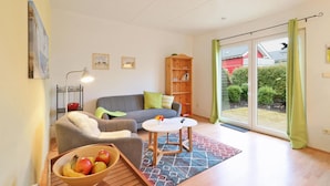 Una sala de estar con un sofá, un sillón y una mesita con un frutero. Hay una estantería y una ventana con cortinas.