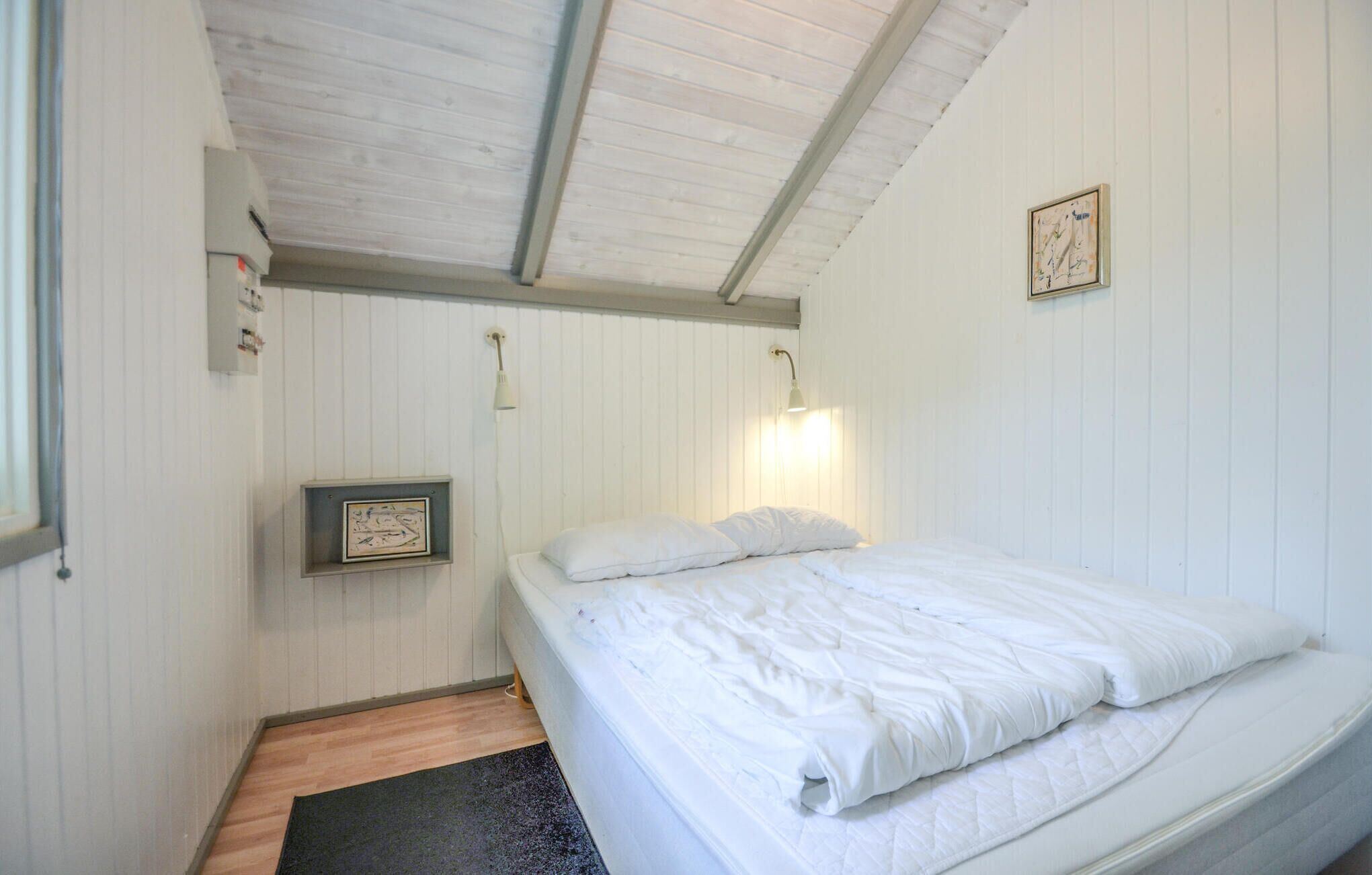 Une petite chambre avec un lit simple, aux murs blancs et au plafond en pente. On y trouve un tableau encadré, une lampe de chevet et une petite étagère sur laquelle est posé un cadre photo.