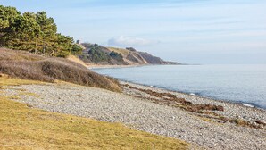 Un paysage côtier avec une plage de galets, des zones herbeuses et une colline lointaine surmontée d’une construction.