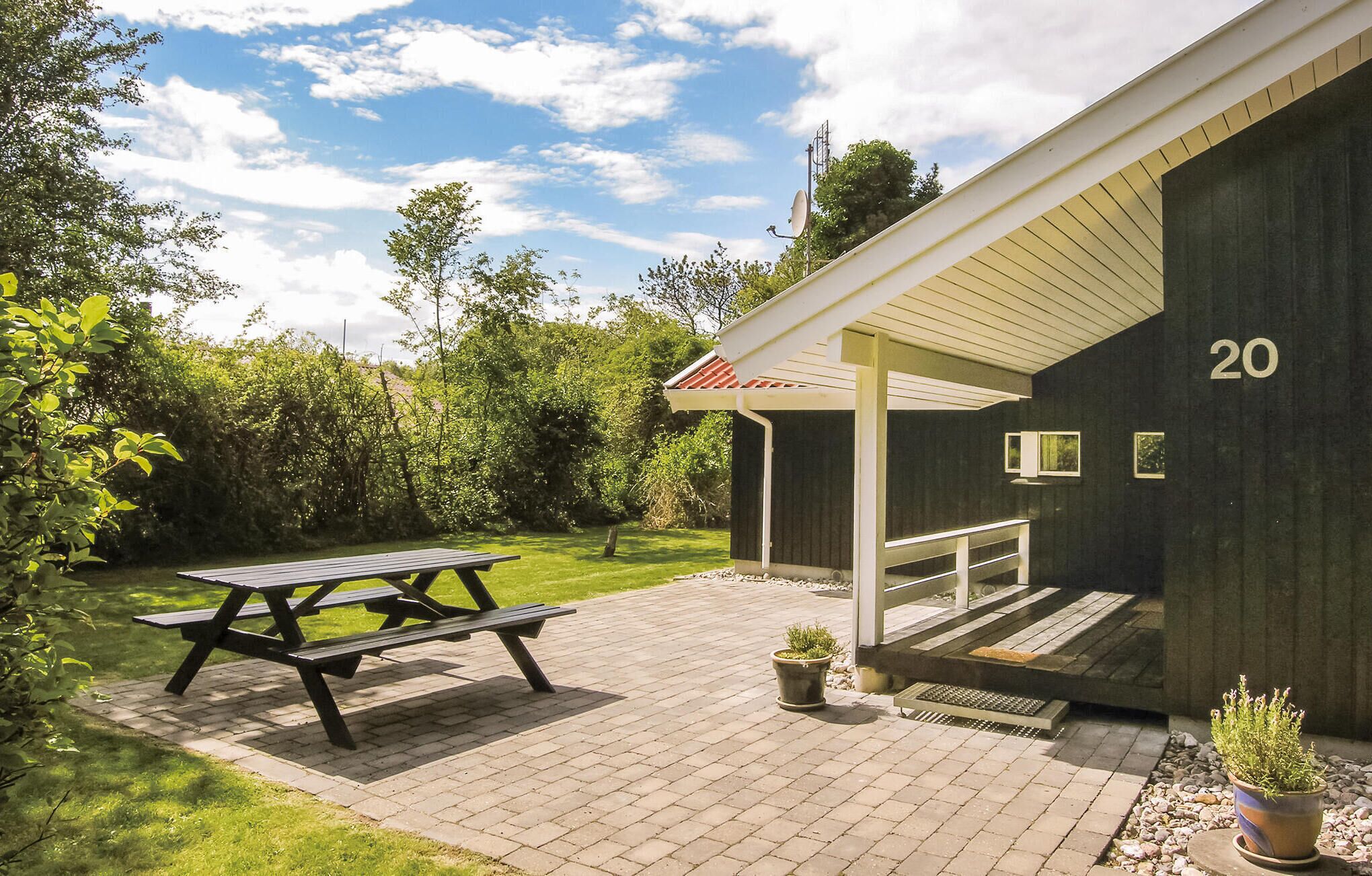 Een overdekt terras met een picknicktafel, een klein gebouw met nummer 20, en een tuin vol potplanten.