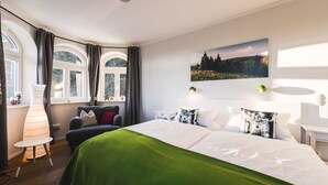 Ein Hotelzimmer mit einem Bett, einem Sessel, einer Lampe, einem Fenster mit Vorhängen und einem Wandbild.