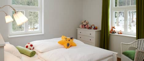Un dormitorio con cama, mesita de noche, lámpara, ventana con cortinas, silla y vista a árboles.
