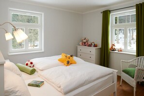 A bedroom with a bed, a nightstand, a lamp, a window with curtains, a chair, and a view of trees.
