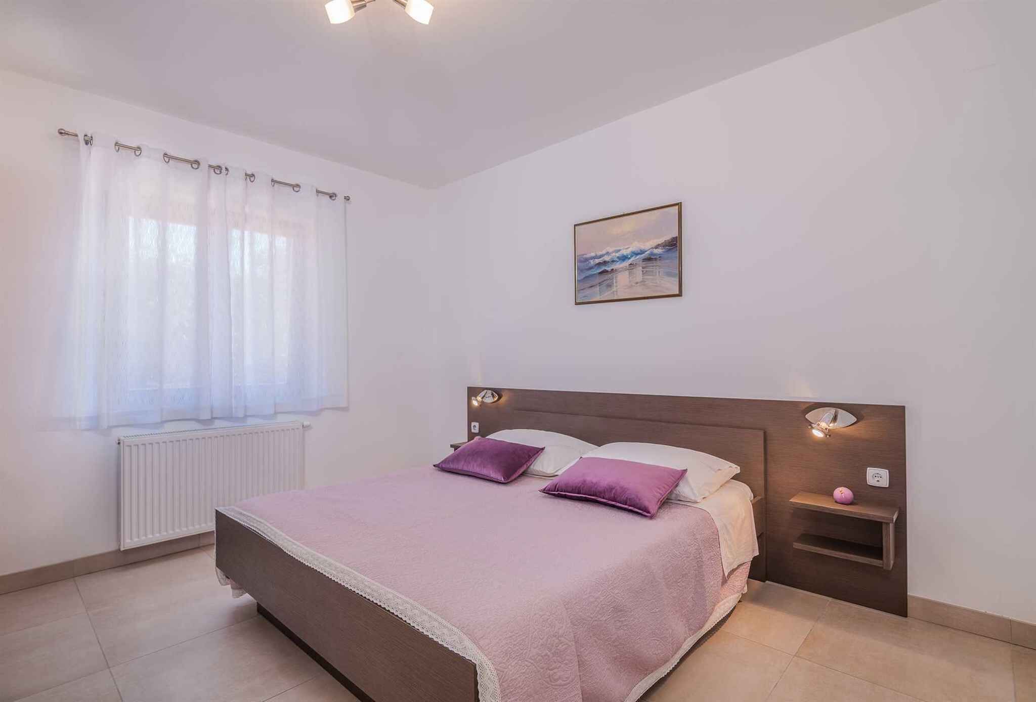 Un dormitorio con cama, mesita de noche, ventana con cortinas, luz de pared e imagen.