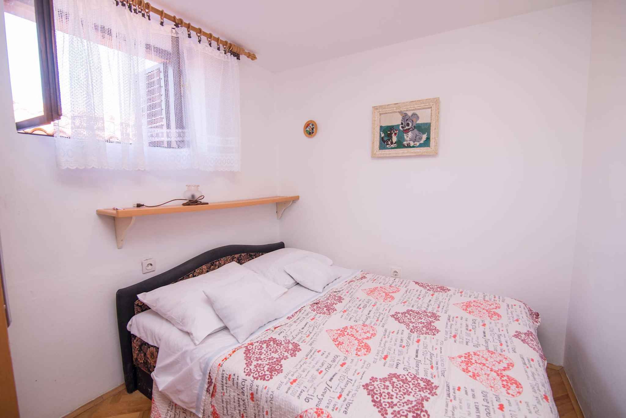 Een slaapkamer met een bed, een raam met kantgordijnen, een klein plankje en een lijst met een foto aan de muur.
