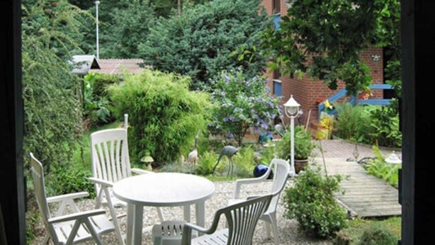 A garden patio with a round table and white chairs, surrounded by greenery and a brick wall in the background.