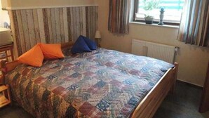 A bedroom with a wooden headboard, a bed with a patterned quilt, two pillows, a nightstand, a lamp, a window with curtains, and a radiator.