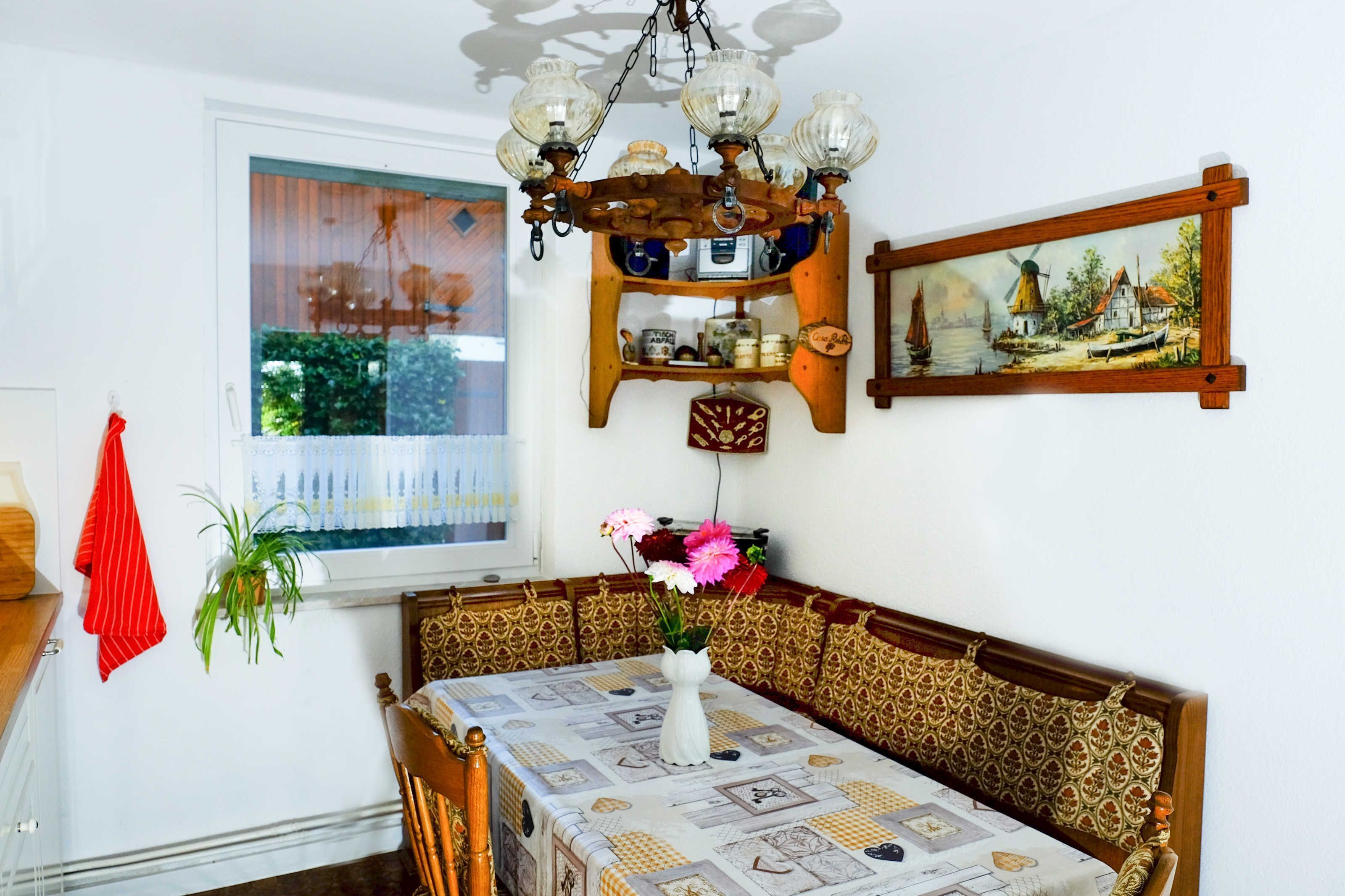 A dining area with a table set for two, a wooden bench, a chandelier, a window with a view, and a wall-mounted shelf with various items.