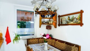 Uma área de jantar com uma mesa posta para dois, um banco de madeira, um candelabro, uma janela com vista e uma estante fixada na parede com diversos objetos.