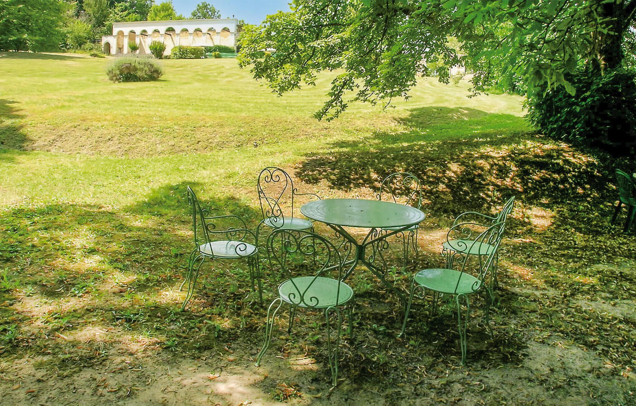 Ein Garten mit einem runden Tisch und vier Stühlen, umgeben von viel Grün und einem Gebäude mit Bögen im Hintergrund.