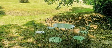 Ein Garten mit einem runden Tisch und vier Stühlen, umgeben von viel Grün und einem Gebäude mit Bögen im Hintergrund.