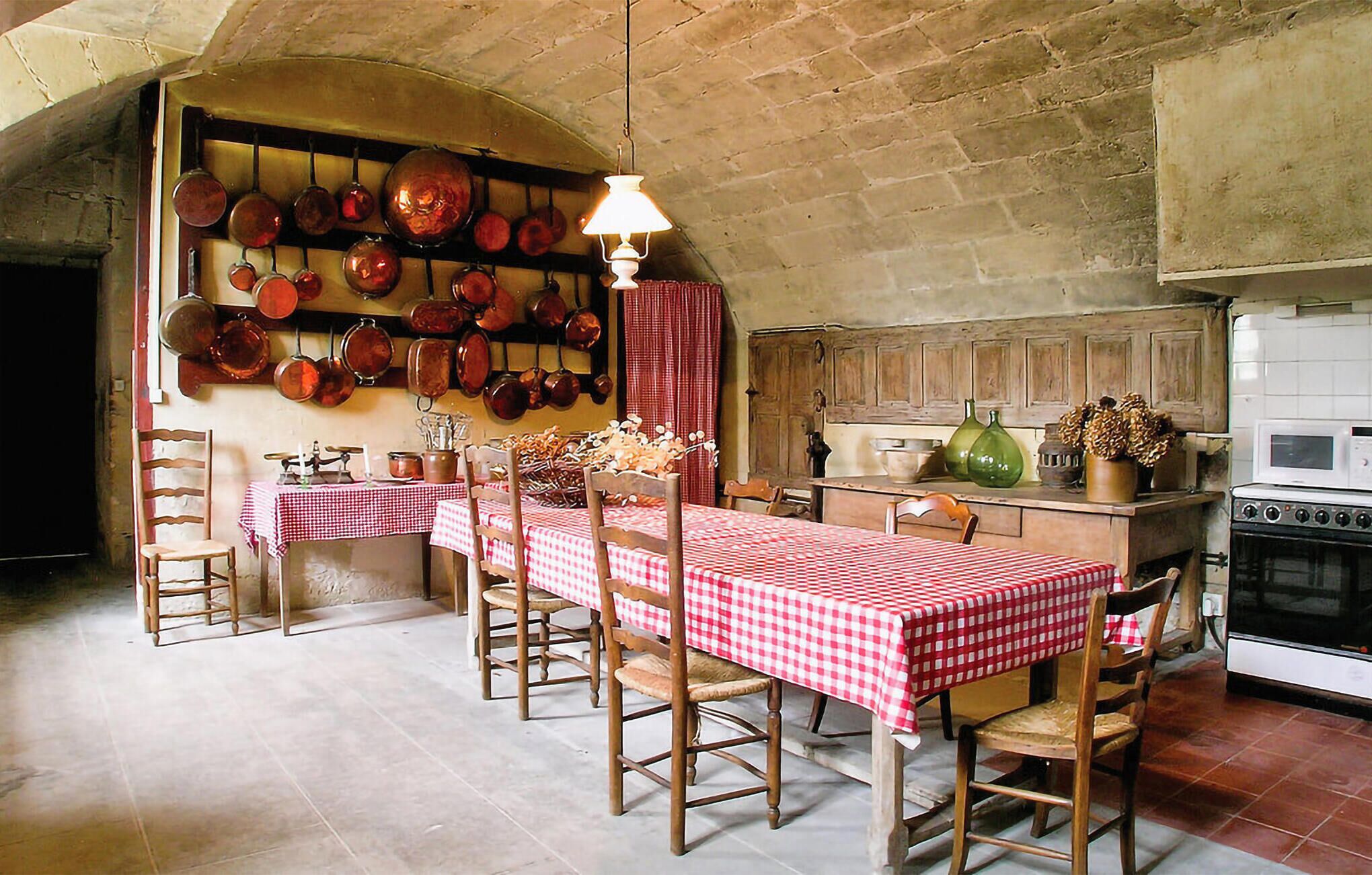 A rustic kitchen with a long dining table, wooden chairs, and a variety of cookware hanging on the wall.