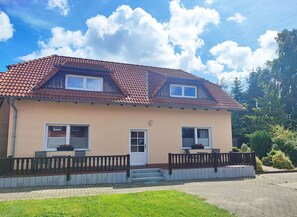 A two-story house with a red-tiled roof, a front porch with a bench, and a small garden area.