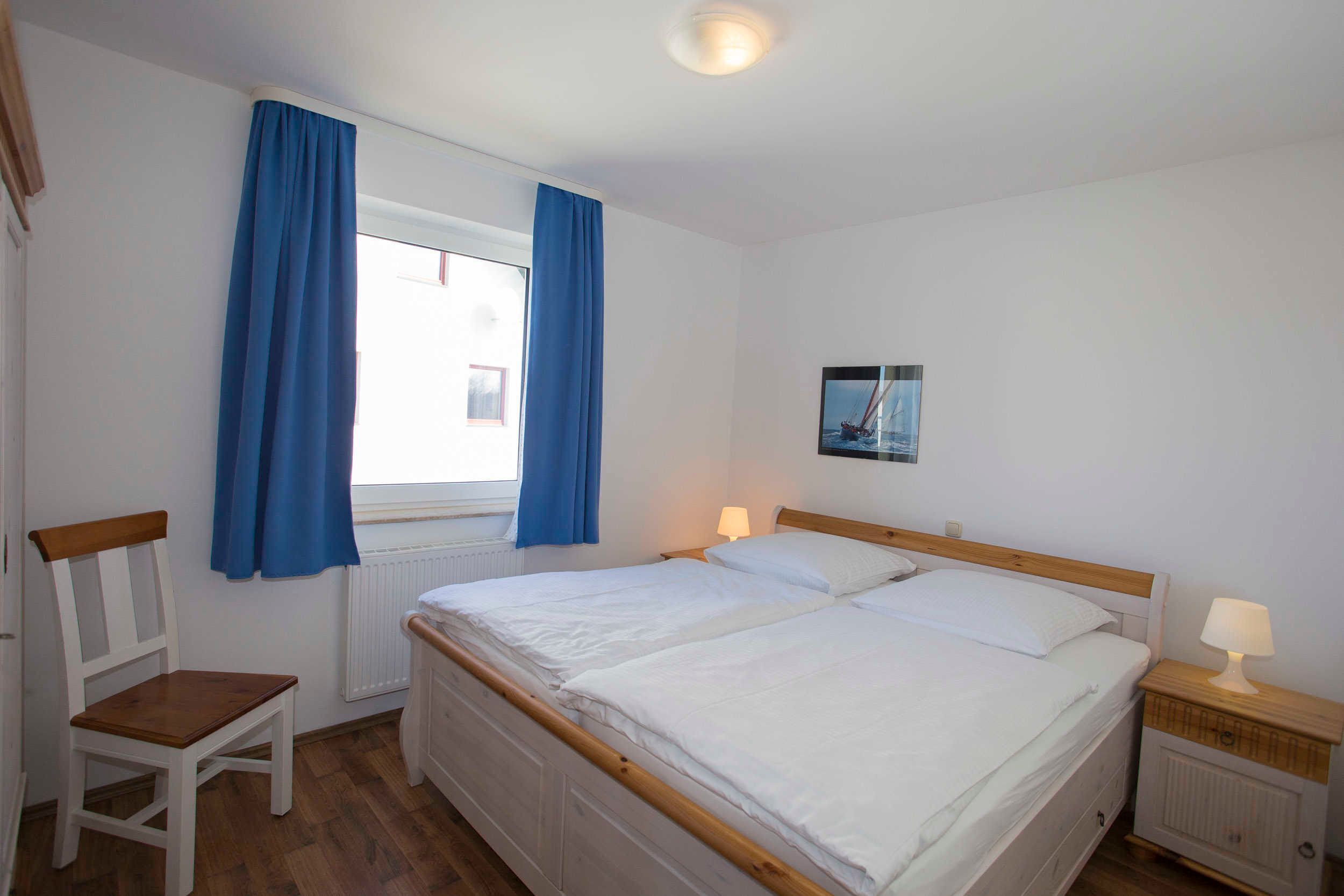 A bedroom with a double bed, a wooden chair, a nightstand with a lamp, and a window with blue curtains.