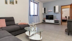 Een moderne woonkamer met een grijze bank, een glazen salontafel, een televisie op een witte kast, en een bed op de achtergrond.