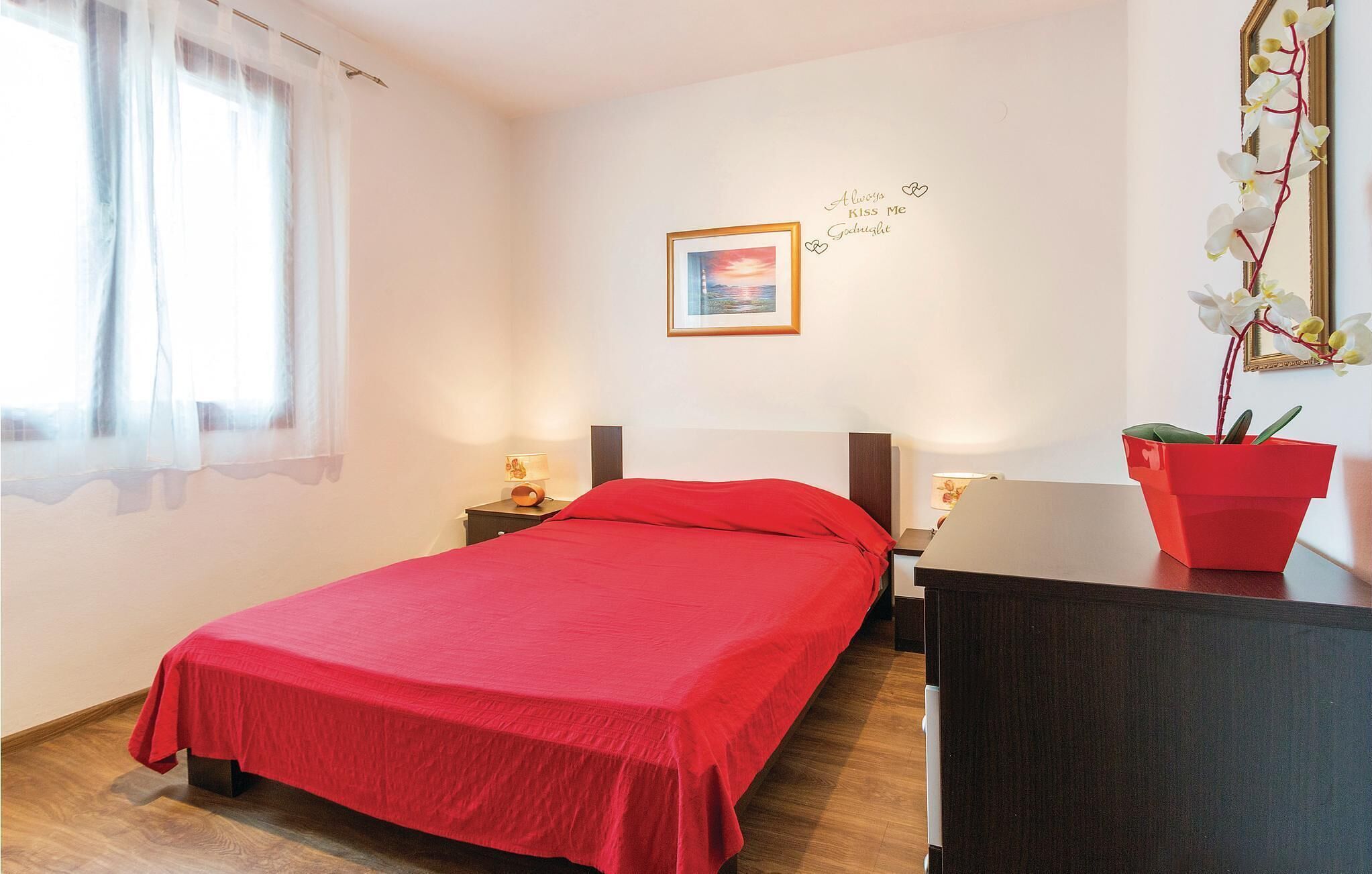 A bedroom with a bed, bedside tables, a desk, a chair, a window with curtains, a wall-mounted picture, and a potted plant.
