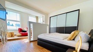 A modern bedroom with a large bed, a bedside table, and a view of a red sofa in the adjacent room. - Villa Anna App. Johann Strauss - Villa Anna App. Johann Strauss (Heringsdorf)