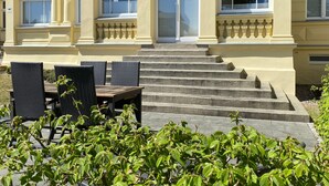 A building with a staircase leading to a balcony, outdoor seating with chairs, and a garden area with plants. - Villa Anna App. Johann Strauss - Villa Anna App. Johann Strauss (Heringsdorf)