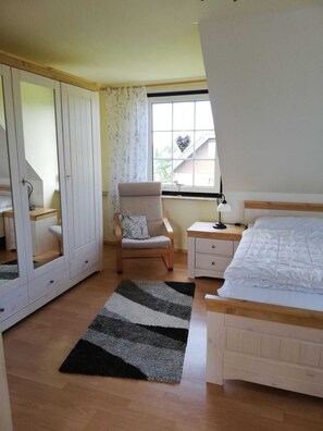 A bedroom with a bed, a chair, a nightstand, a desk, and a window with a view of a house.