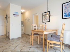 A dining area with a wooden table and chairs, a kitchen in the background, and a wall with a framed picture and a decorative sign.