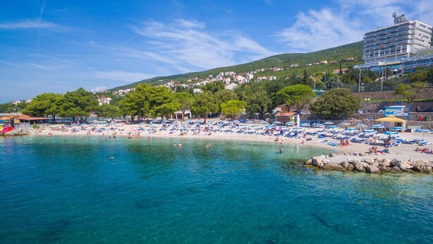 Ein Strand mit klarem, blauem Wasser, vielen Liegestühlen und einem Gebäude im Hintergrund.