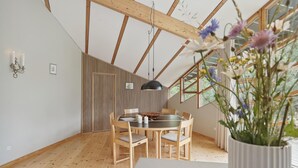 Un espace repas avec une table et des chaises en bois, un luminaire suspendu et une vue sur l’extérieur grâce à de grandes fenêtres.