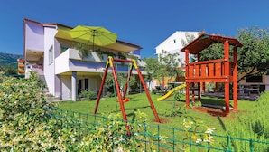 Een speeltuin met schommels en een glijbaan, een groene parasol, en een rode speelhoes voor een gebouw met twee verdiepingen, een witte gevel en een rood dak.