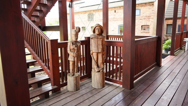 Een houten terras met twee houten sculpturen, een houten trap en een bakstenen gebouw op de achtergrond.