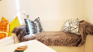 Een woonkamer met een witte bank, een luipaardprint plaid en gestreepte kussens. Voor de bank staat een klein wit tafeltje met een groene mat en twee bruine objecten.