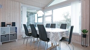 A modern dining area with a round table, black chairs, and a white tablecloth. There are speakers on a shelf, a plant on a stand, and a window with a view of trees. - Gorgeous home in Løkken with sauna (Løkken)