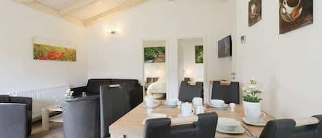 A dining area with a wooden table set for four, surrounded by black chairs. There is a sofa and a small table with a vase of flowers. The room has white walls, wooden beams on the ceiling, and artwork on the walls.