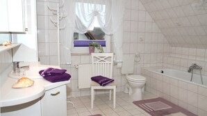 Une salle de bain avec un lavabo, des toilettes et une baignoire, tous de couleur blanche. On y trouve une fenêtre avec un rideau, un radiateur et une serviette violette posée sur une chaise.
