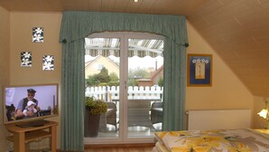 Une chambre avec une télévision, un lit avec une literie à motifs floraux, une porte-fenêtre coulissante donnant sur un balcon aménagé avec du mobilier d’extérieur, et une clôture blanche en bois.
