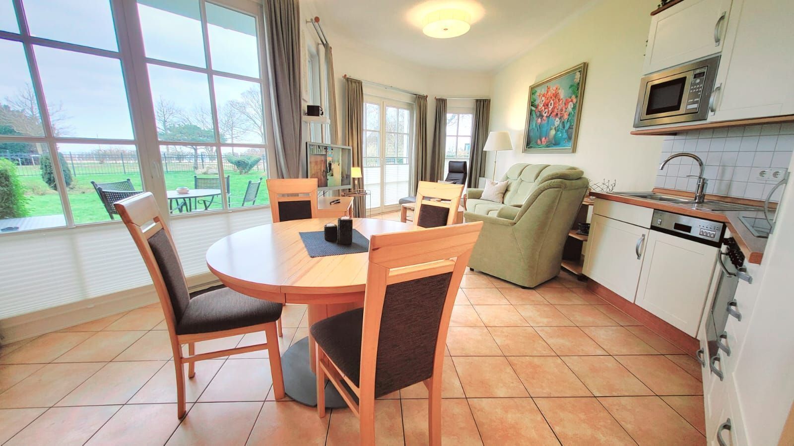 A dining area with a round table and chairs, a sofa, a kitchen with a microwave and sink, and large windows with curtains.