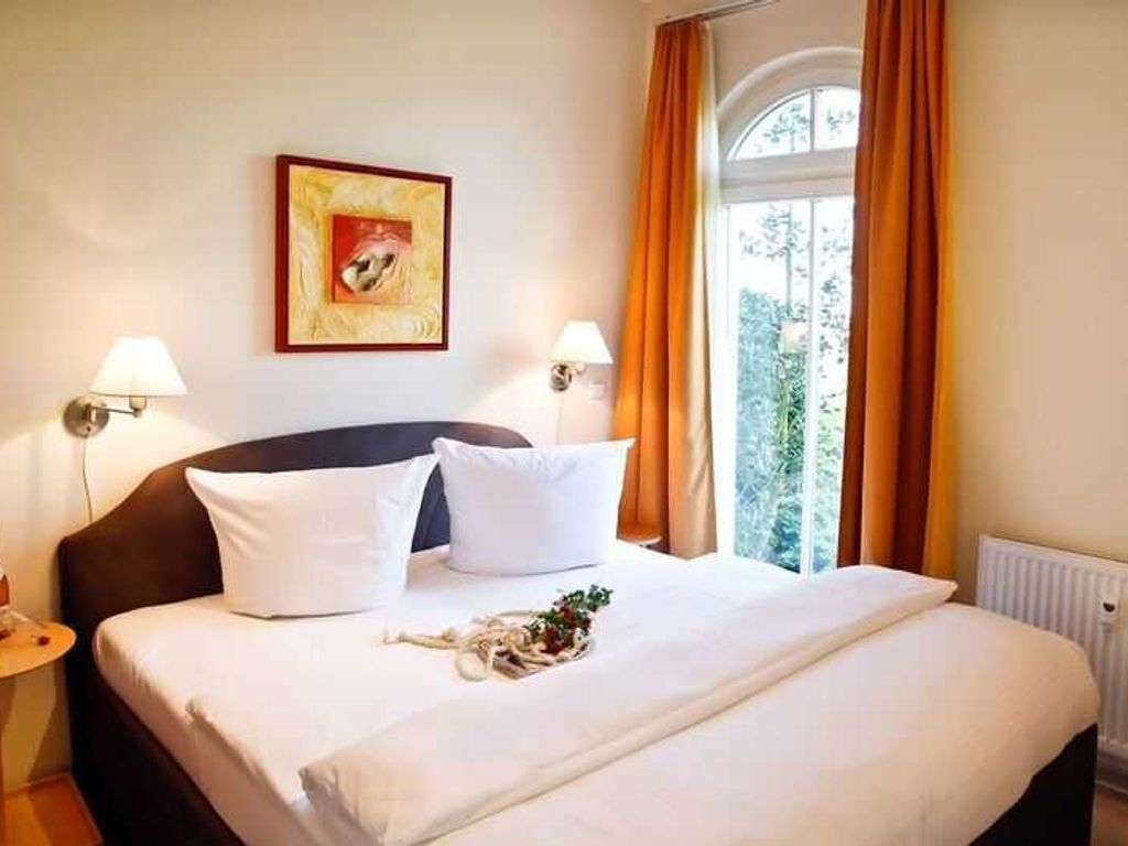 Ein Hotelzimmer mit einem Bett, zwei Nachttischlampen, einem gerahmten Bild an der Wand und einem Fenster mit Vorhängen.