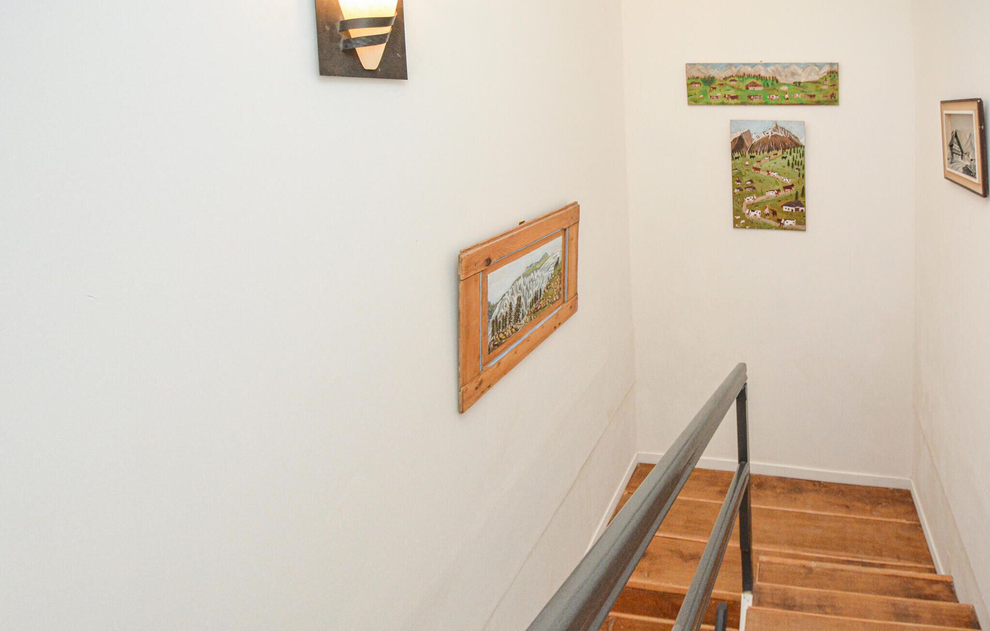 A staircase with wooden steps and a metal handrail, flanked by white walls with framed artwork.