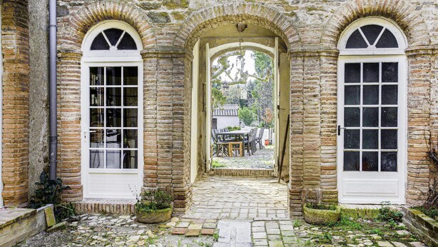 Un bâtiment en pierre doté de trois fenêtres en arc de cercle et d’une porte d’entrée centrale qui donne sur une cour intérieure avec une table et des chaises.