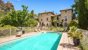 Une piscine avec des chaises longues, un bâtiment en pierre doté d’un toit en tuiles et un jardin clos.