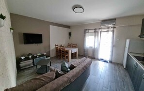 Een moderne woonkamer met een bank, een aan de muur bevestigde televisie, een eettafel met stoelen en een keuken met een koelkast.
