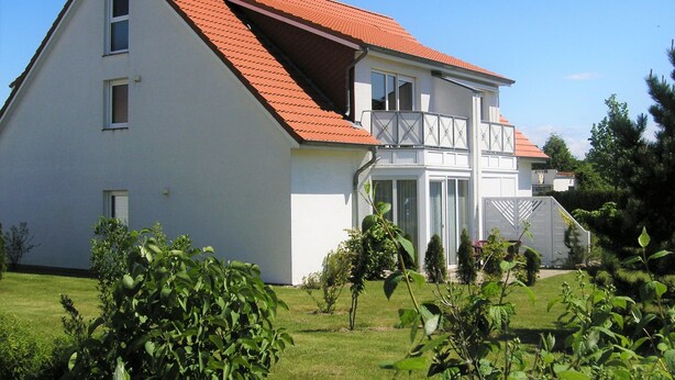 Une maison à deux étages, avec un toit rouge, des murs extérieurs blancs et un balcon doté d’une balustrade.
